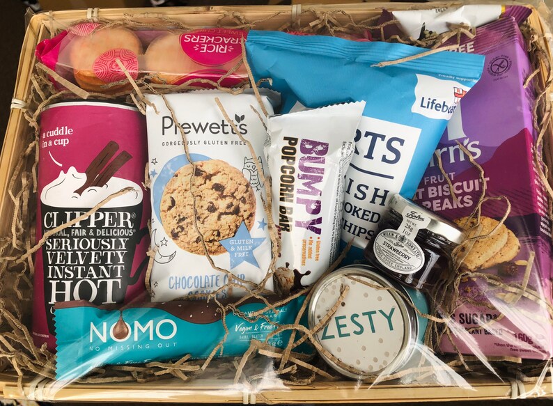 May include: A gift basket filled with various snacks and treats, including a bag of popcorn, a bag of chips, a jar of jam, a box of cookies, a chocolate bar, and a box of instant hot chocolate. The basket is lined with brown paper shreds and has a clear plastic wrap over the top.