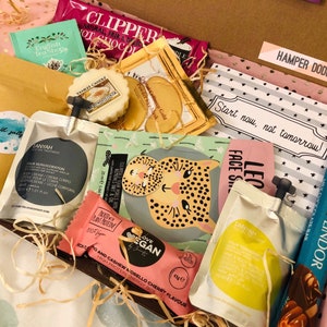 May include: A gift box filled with various self-care items. The box contains tea bags, a Yankee Candle, face masks, bath salts, chocolate, and a vegan energy bar. The box is lined with pink and gold polka dot paper and includes a card that says "Start now, not tomorrow!"