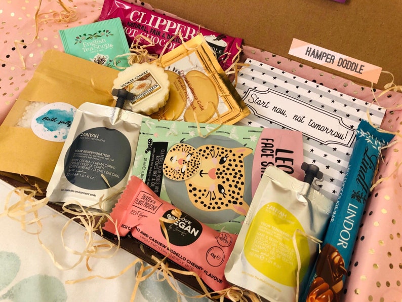 May include: A gift box filled with various self-care items. The box contains tea bags, a Yankee Candle, face masks, bath salts, chocolate, and a vegan energy bar. The box is lined with pink and gold polka dot paper and includes a card that says "Start now, not tomorrow!"