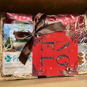 May include: A brown wicker basket filled with a variety of Christmas themed gifts, including cookies, a card, and a pair of socks. The basket is wrapped with a brown ribbon and a red and white patterned paper.