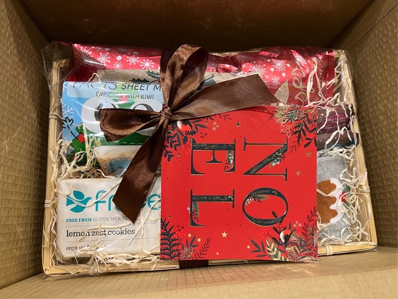May include: A brown wicker basket filled with a variety of Christmas themed gifts, including cookies, a card, and a pair of socks. The basket is wrapped with a brown ribbon and a red and white patterned paper.