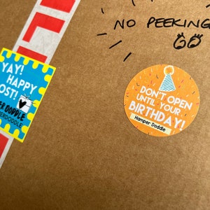 May include: A brown cardboard box with a yellow and blue sticker that says "YAY! HAPPY POST!" and a yellow sticker with confetti and the text "DON'T OPEN UNTIL YOUR BIRTHDAY!"  The box also has a red and white label that says "LIF" and handwritten text that says "NO PEEKING!"
