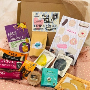 May include: A gift box filled with self-care items, including a pineapple face mask, snacks, tea bags, and a notepad. The box also contains a bar of chocolate, eye patches, and a candle. The text on the box reads "Press Pause".