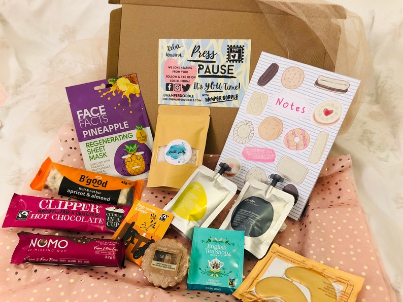 May include: A gift box filled with self-care items, including a pineapple face mask, snacks, tea bags, and a notepad. The box also contains a bar of chocolate, eye patches, and a candle. The text on the box reads "Press Pause".