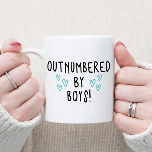 May include: White ceramic mug with black text that reads "Outnumbered by Boys!" with blue hearts around the text.