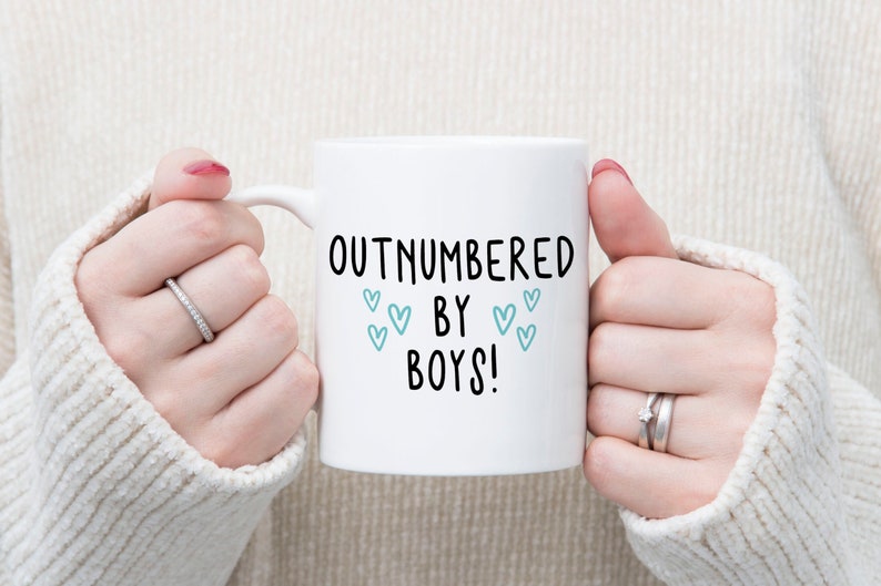 May include: White ceramic mug with black text that reads "Outnumbered by Boys!" with blue hearts around the text.