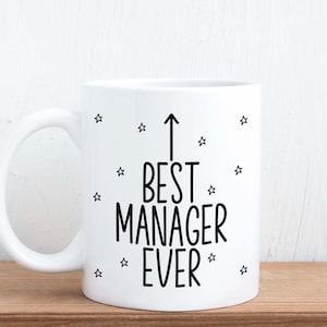 May include: White ceramic mug with the text "BEST MANAGER EVER" in black, with an arrow pointing upwards. Small black star shapes are scattered around the text. The mug has a curved handle.