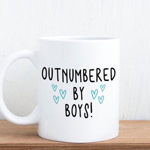 May include: White ceramic coffee mug with black text that reads "Outnumbered by Boys!" with blue heart illustrations.