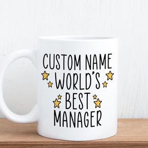 May include: A white ceramic mug featuring black text that reads CUSTOM NAME WORLD'S BEST MANAGER, accented by six small yellow stars arranged around the lettering. The mug has a standard handle on the left side and a smooth, glossy finish.