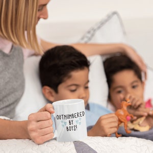 May include: A white ceramic mug with the text "Outnumbered by Boys!" in black lettering and small blue hearts. The mug is being held by a person's hand. Two children are sitting in the background.