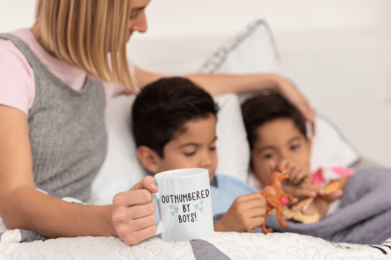 May include: A white ceramic mug with the text "Outnumbered by Boys!" in black lettering and small blue hearts. The mug is being held by a person's hand. Two children are sitting in the background.