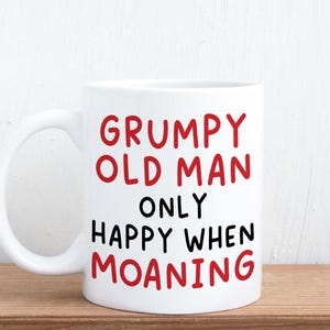 May include: White ceramic mug with the text "GRUMPY OLD MAN ONLY HAPPY WHEN MOANING" in red and black lettering. The mug has a curved handle and is set against a light wood surface and a white background.