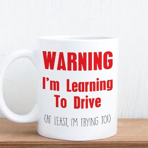 May include: White ceramic mug with the text "WARNING I'm Learning To Drive (CAT LEAST, I'M TRYING TO)" in red. The mug is on a wooden surface against a white background. The mug has a curved handle.