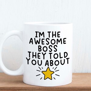 May include: White ceramic mug featuring the text "I'M THE AWESOME BOSS THEY TOLD YOU ABOUT" in black, with a yellow star graphic. The mug has a curved handle and is set against a light wood surface and white background.