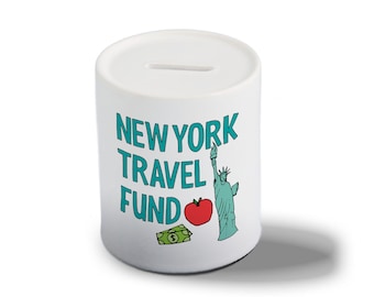 May include: White ceramic piggy bank with a slot on top. The bank has a blue and green design with the text "NEW YORK TRAVEL FUND" and an illustration of the Statue of Liberty, an apple, and a dollar bill.