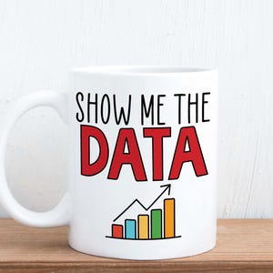 May include: White ceramic mug with the text "SHOW ME THE DATA" in black and red. Below the text is a colourful bar graph with an upward trending arrow. The mug has a curved handle.