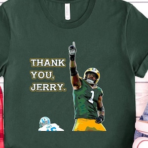 May include: Forest green t-shirt featuring a football player in a green and yellow uniform with the number 1, raising his index finger. The text "THANK YOU, JERRY." is printed on the shirt. A football helmet is on the ground.