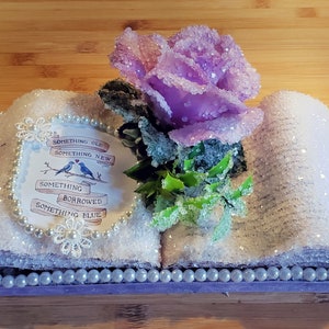 May include: A white book with a purple rose and a pearl trim. The book has a white background with the words "Something Old, Something New, Something Borrowed, Something Blue" written on it. Two blue birds are perched on a branch above the words.