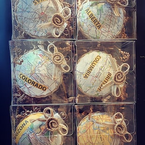Puede incluir: Seis cajas de plástico transparente que contienen cada una un adorno redondo de mapa con un lazo de arpillera y una pequeña flor de arpillera. Los adornos están decorados con mapas de diferentes estados y provincias, incluyendo Texas, Alberta, Columbia Británica, Colorado y Arizona.