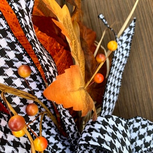 K&ouml;nnte beinhalten: Ein schwarz-wei&szlig;es Hahnentrittband mit orangefarbenen und roten Beeren und braunen Bl&auml;ttern. Das Band ist zu einer Schleife gebunden und Teil eines Herbstkranzes.