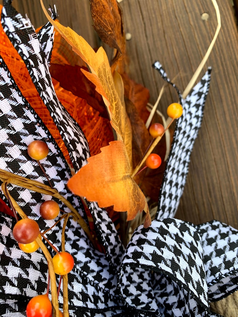 K&ouml;nnte beinhalten: Ein schwarz-wei&szlig;es Hahnentrittband mit orangefarbenen und roten Beeren und braunen Bl&auml;ttern. Das Band ist zu einer Schleife gebunden und Teil eines Herbstkranzes.