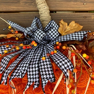 K&ouml;nnte beinhalten: Eine Schleife aus schwarz-wei&szlig;em kariertem Band mit einem Jutestiel, einem orangefarbenen Netz-Kranz und herbstfarbenen Beeren.