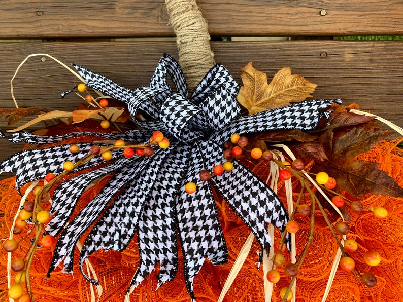 K&ouml;nnte beinhalten: Eine Schleife aus schwarz-wei&szlig;em kariertem Band mit einem Jutestiel, einem orangefarbenen Netz-Kranz und herbstfarbenen Beeren.