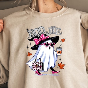 May include: A beige crewneck sweatshirt featuring a graphic of a ghost wearing a black witch hat with a pink polka dot bow, pink sunglasses, and pink heels. The ghost holds a coffee cup and a leopard print handbag. The text BOO JIE is above.