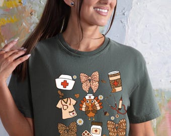 May include: A dark green t-shirt featuring a collection of fall-themed medical and nurse-related illustrations. The design includes a nurse's cap, bow, coffee cup, turkey, and medical tools. The shirt is made of a soft material.