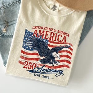 Op de afbeelding: Crèmekleurig T-shirt met een afbeelding van de Amerikaanse vlag en een adelaar, met de tekst "UNITED STATES OF AMERICA AMERICA 250th Anniversary 1776-2026." Het shirt wordt gecombineerd met een lichtblauwe jeans en een beige hoed.