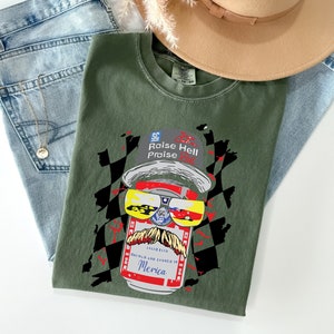 May include: Green t-shirt with a graphic of a can of beer wearing sunglasses and a hat that says "SC Raise Hell Praise". The can has a mustache and the text "Born and Canned in 'Merica".