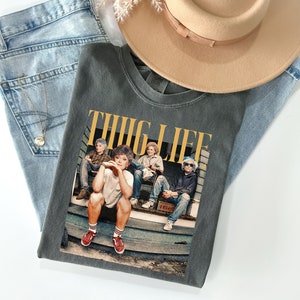 May include: A dark gray t-shirt with a graphic print of four people sitting on a porch. The text "THUG LIFE" is printed in yellow letters above the image.