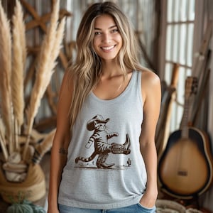May include: A light gray tank top featuring a brown graphic of a cat wearing a cowboy hat and boots. The cat is walking with one leg raised. A cactus is also in the graphic.