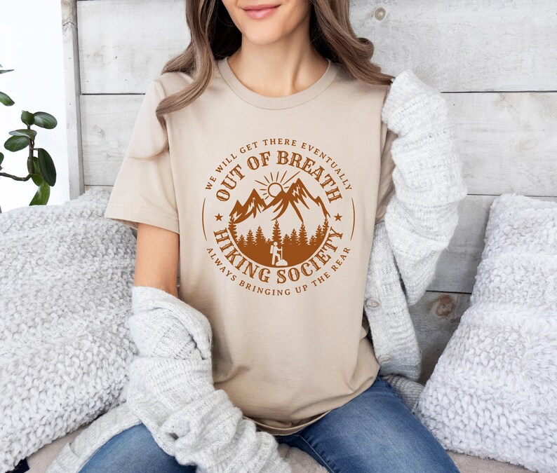 Może przedstawiać: Beżowa koszulka z brązową, okrągłą grafiką. Grafika przedstawia g&oacute;ry, drzewa i tekst "WE WILL GET THERE EVENTUALLY OUT OF BREATH HIKING SOCIETY ALWAYS BRINGING UP THE REAR".