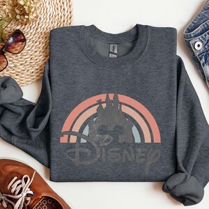 May include: Dark gray sweatshirt featuring a retro rainbow design with a castle silhouette and the word "Disney" in a distressed font. The sweatshirt is a crew neck style and appears to be made of a soft material. The design incorporates shades of orange, pink, and blue.