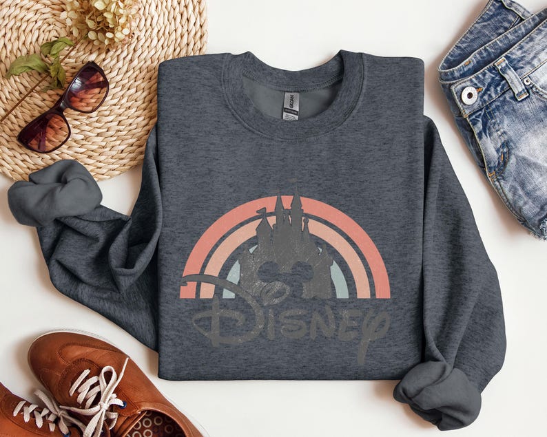 May include: Dark gray sweatshirt featuring a retro rainbow design with a castle silhouette and the word "Disney" in a distressed font. The sweatshirt is a crew neck style and appears to be made of a soft material. The design incorporates shades of orange, pink, and blue.