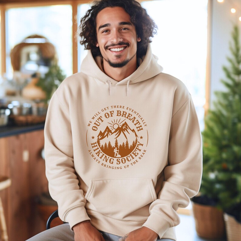 Może przedstawiać: Beżowa bluza z kapturem z przednią kieszenią i kapturem ze sznurkiem. Bluza z kapturem ma brązową grafikę z napisem "OUT OF BREATH HIKING SOCIETY" oraz wzorem g&oacute;r i drzew. Zawiera r&oacute;wnież tekst "WE WILL GET THERE EVENTUALLY" i "ALWAYS BRINGING UP THE REAR".