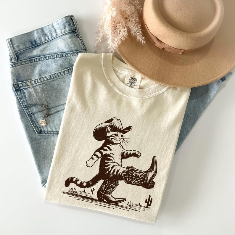 May include: Cream-colored t-shirt featuring a cartoon cat wearing a cowboy hat and boots. The cat is walking on a line with cacti. The shirt is paired with light blue jeans, a necklace, and a tan hat.