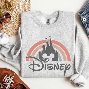 May include: A grey sweatshirt featuring a vintage-style rainbow and a silhouette of Cinderella's castle with Mickey Mouse ears. The text "DISNEY" is printed below the castle.