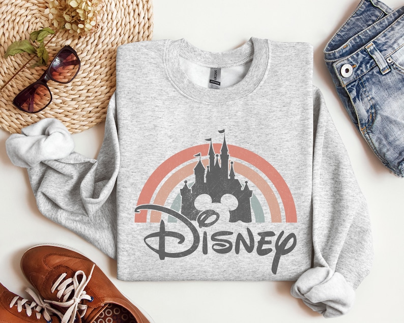 May include: A grey sweatshirt featuring a vintage-style rainbow and a silhouette of Cinderella's castle with Mickey Mouse ears. The text "DISNEY" is printed below the castle.