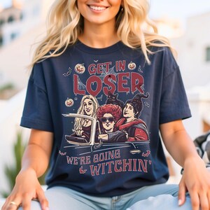 May include: A navy blue t-shirt with a graphic of three witches driving a car. The text on the shirt reads "Get in Loser, We're Going Witchin'"