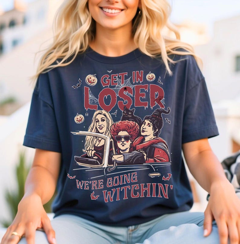 May include: A navy blue t-shirt with a graphic of three witches driving a car. The text on the shirt reads "Get in Loser, We're Going Witchin'"