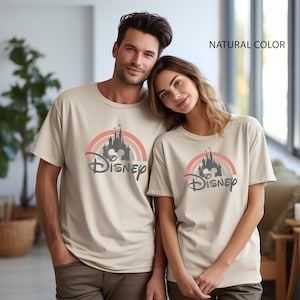 May include: Two beige t-shirts with a Disney castle design. The design features a rainbow, a castle silhouette, and the Disney logo. The text "NATURAL COLOR" is visible in the top right corner.