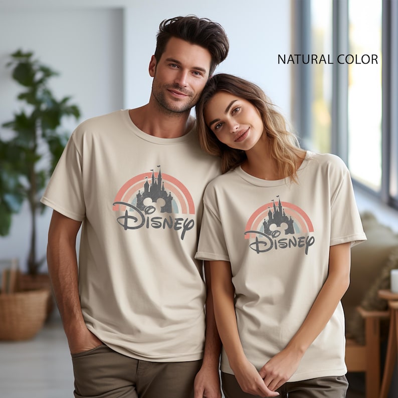 May include: Two beige t-shirts with a Disney castle design. The design features a rainbow, a castle silhouette, and the Disney logo. The text "NATURAL COLOR" is visible in the top right corner.