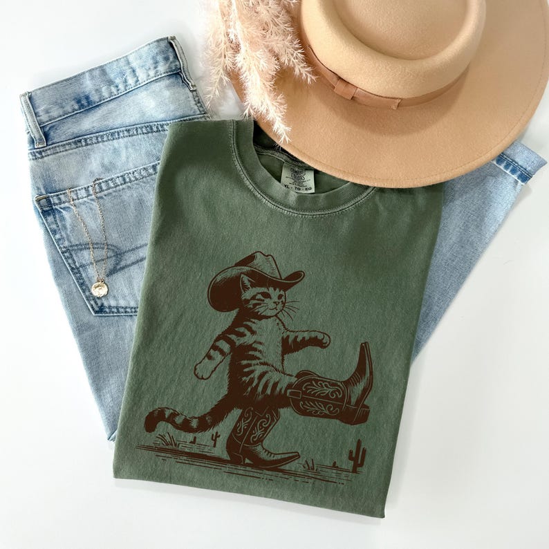 May include: An olive green t-shirt featuring a brown illustration of a cat wearing a cowboy hat and boots. The shirt is paired with light wash denim jeans and a tan hat. A gold necklace is visible on the jeans.