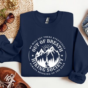 Może przedstawiać: Granatowa bluza z białym nadrukiem. Okrągła grafika zawiera tekst "We Will Get There Eventually Out of Breath Hiking Society Always Bringing Up The Rear" z motywem g&oacute;r i turysty.