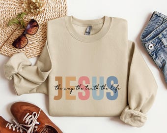 May include: A beige crewneck sweatshirt with the word "JESUS" in brown, peach, pink, and blue block letters. Below the word "JESUS" is the phrase "the way the truth the life" in a black script font. Sunglasses, shoes, and jeans are also in the image.