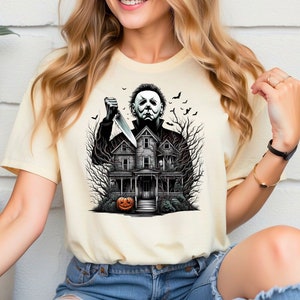 May include: A cream-colored t-shirt with a graphic print of a horror movie character holding a knife in front of a house with a jack-o-lantern in front of it.