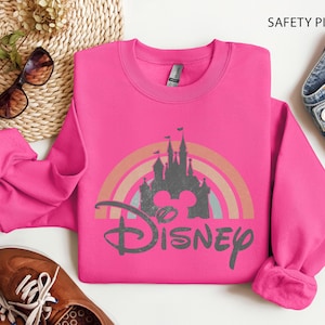 May include: A safety pink sweatshirt with a Disney graphic. The graphic features a castle silhouette, a rainbow, and the Disney logo. The sweatshirt is a crew neck style.