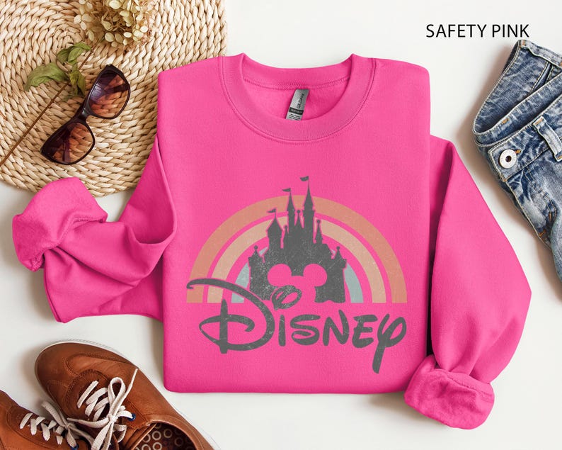 May include: A safety pink sweatshirt with a Disney graphic. The graphic features a castle silhouette, a rainbow, and the Disney logo. The sweatshirt is a crew neck style.